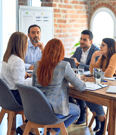 A group of five people having a meeting around a wooden table in a room with brick walls and large arched windows.