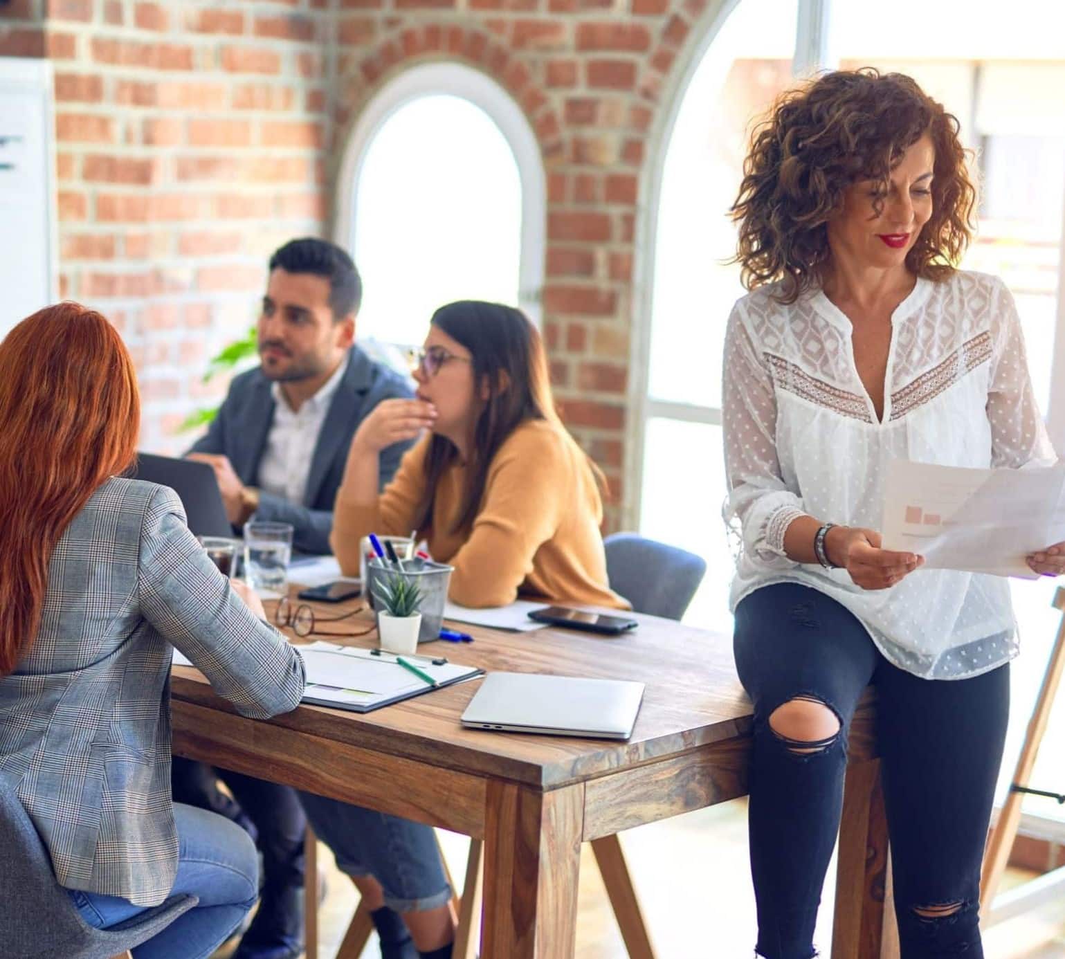 A group of people having a meeting around a wooden table in a room with brick walls and large arched windows.